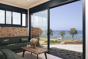 A modern living room with a stone wall, a green sofa, a wooden coffee table, and a view of the sea. - Pelagos SeaSide Suites (Hersonissos)