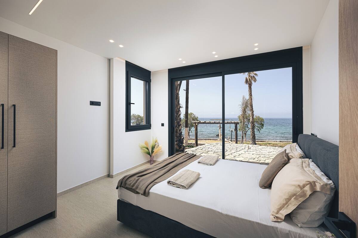 A bedroom with a large bed, a wardrobe, a view of the sea, and a balcony with a pergola.