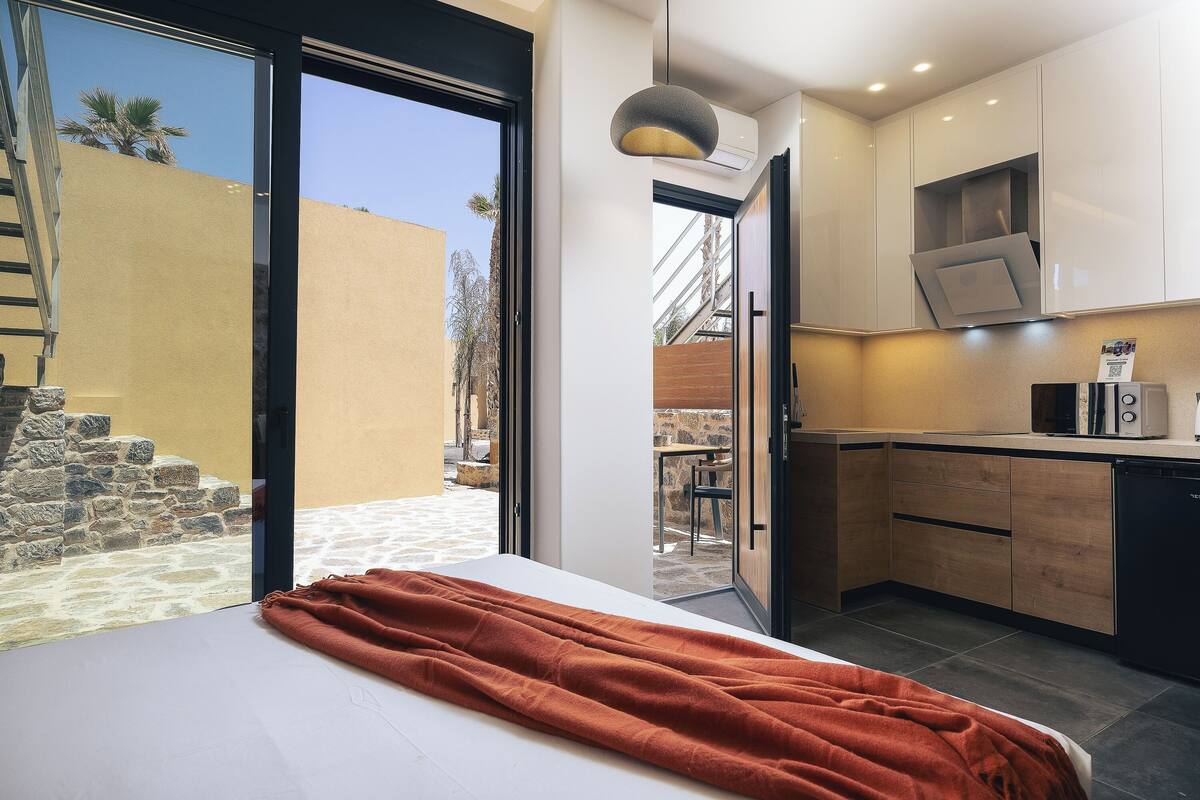 A bedroom with a bed, a red blanket, a sliding glass door leading to a patio, and a kitchen with wooden cabinets and a microwave.