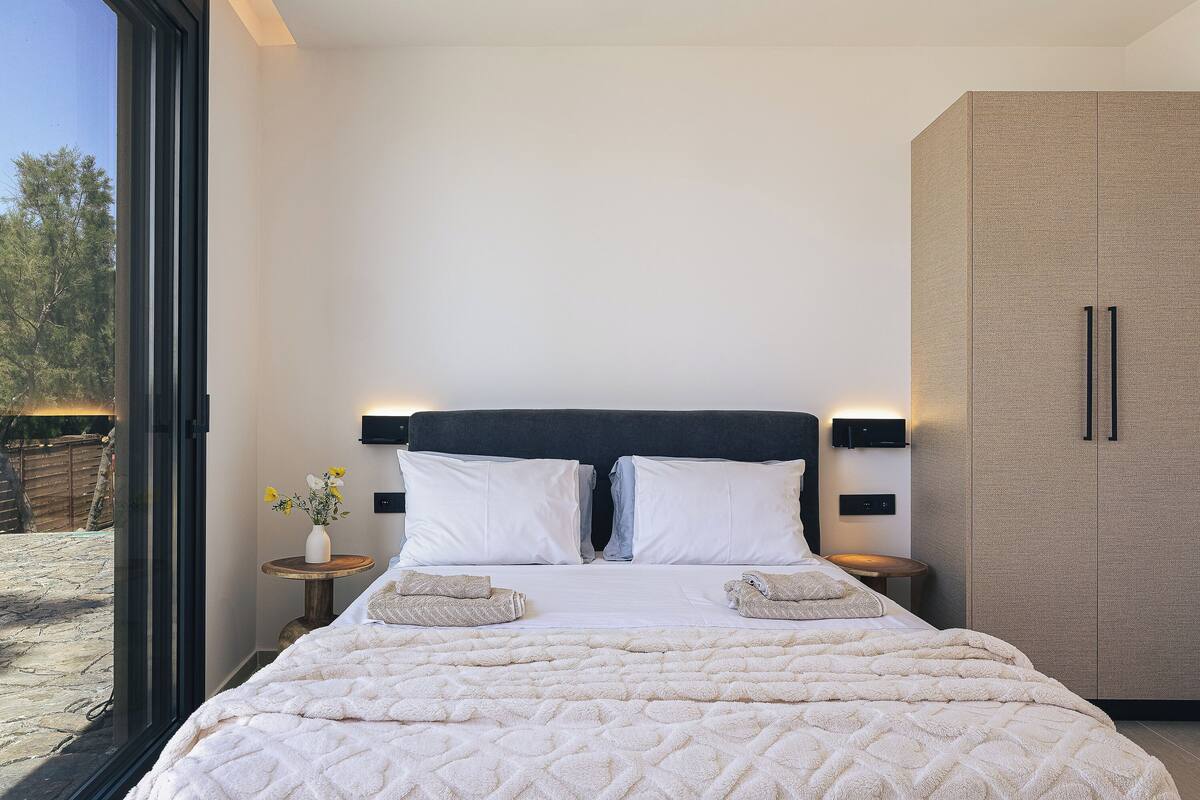 A bedroom with a large bed, bedside tables, a wardrobe, and a view through a glass door.
