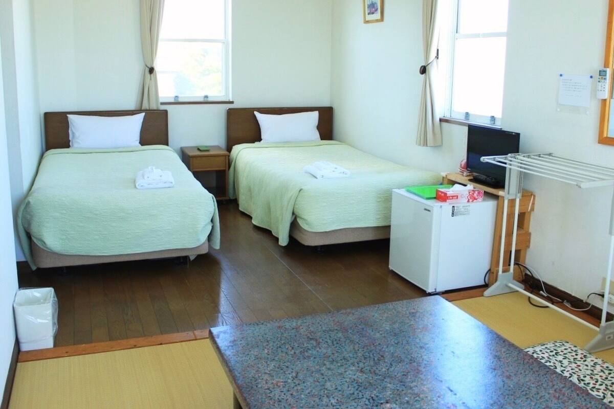 A hotel room with two beds, a small refrigerator, a desk with a computer, and a window with curtains.