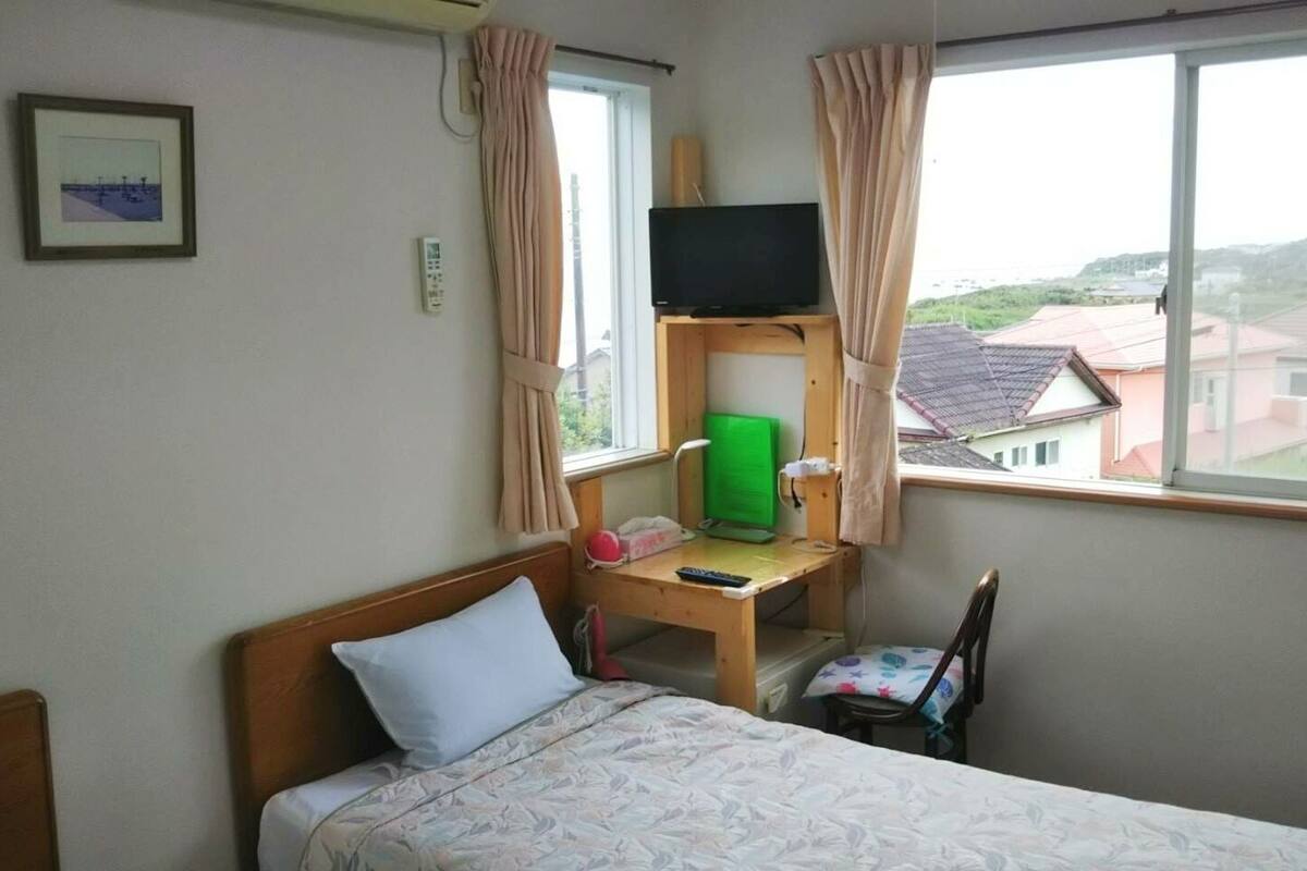 A hotel room with a bed, a desk with a computer and a TV, a chair, and a window with a view of buildings.