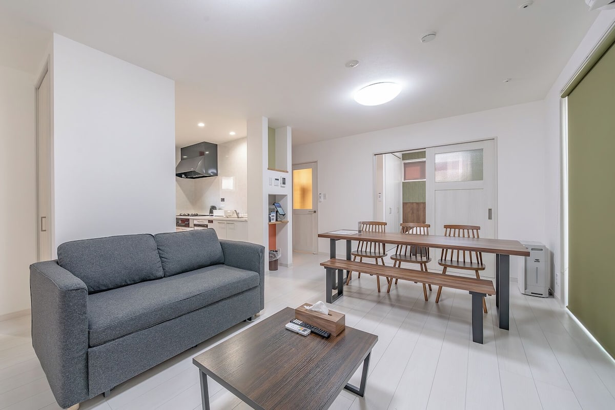 Un appartement moderne avec un canapé gris, une table à manger en bois avec des chaises, un coin cuisine et une petite table basse avec une télécommande.