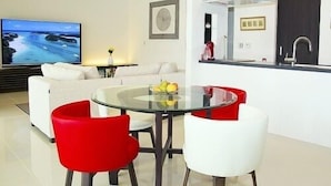 A modern living room with a sofa, a glass table with chairs, a TV, and a kitchen area with a sink. - The Seven Stars Resort (Ishigaki)