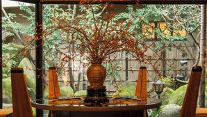 A round dining table with a vase of branches and leaves, surrounded by wooden chairs.