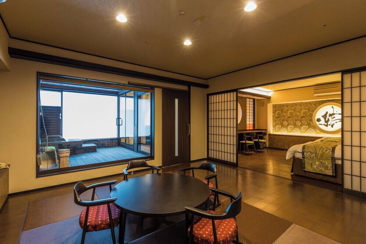 Una habitación de hotel moderna con un amplio ventanal corredero que da a un balcón, una mesa de comedor redonda con sillas, una cama con cabecera de estilo japonés tradicional y una pared decorativa con un emblema circular.