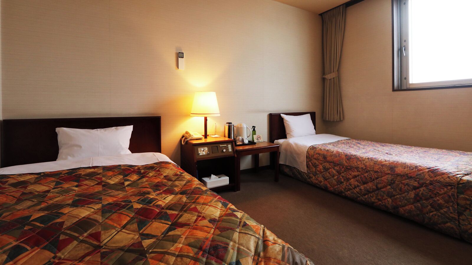 A hotel room with two beds, a nightstand with a lamp, a telephone, and a small table with a bottle and a tissue box.