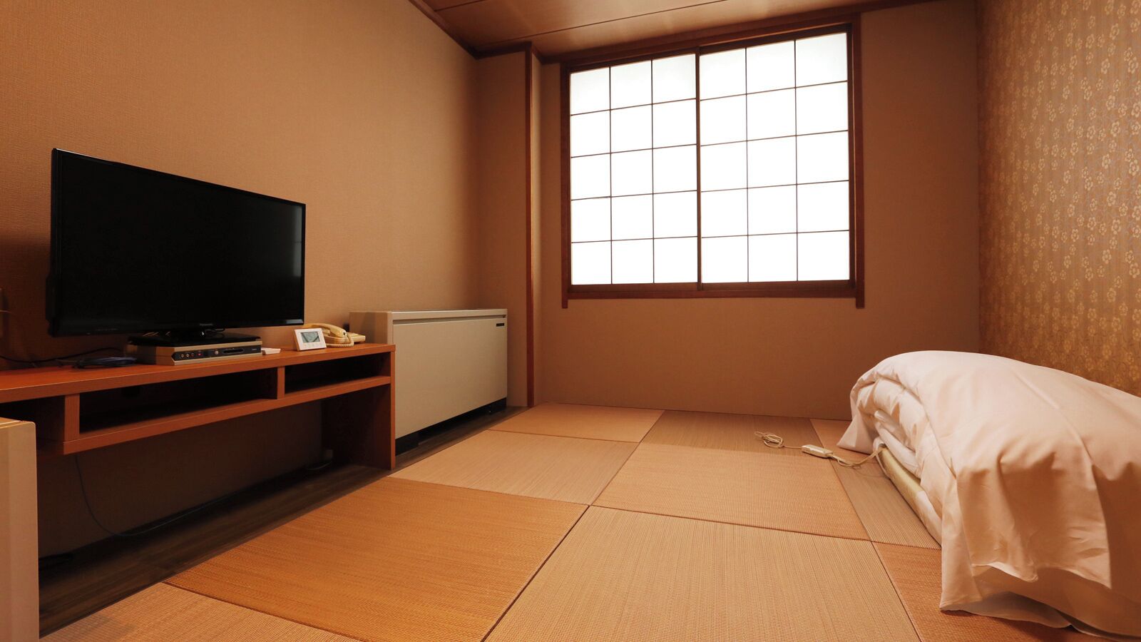 A room with tatami flooring, a television on a wooden stand, a bed with white bedding, and a window with a grid pattern.