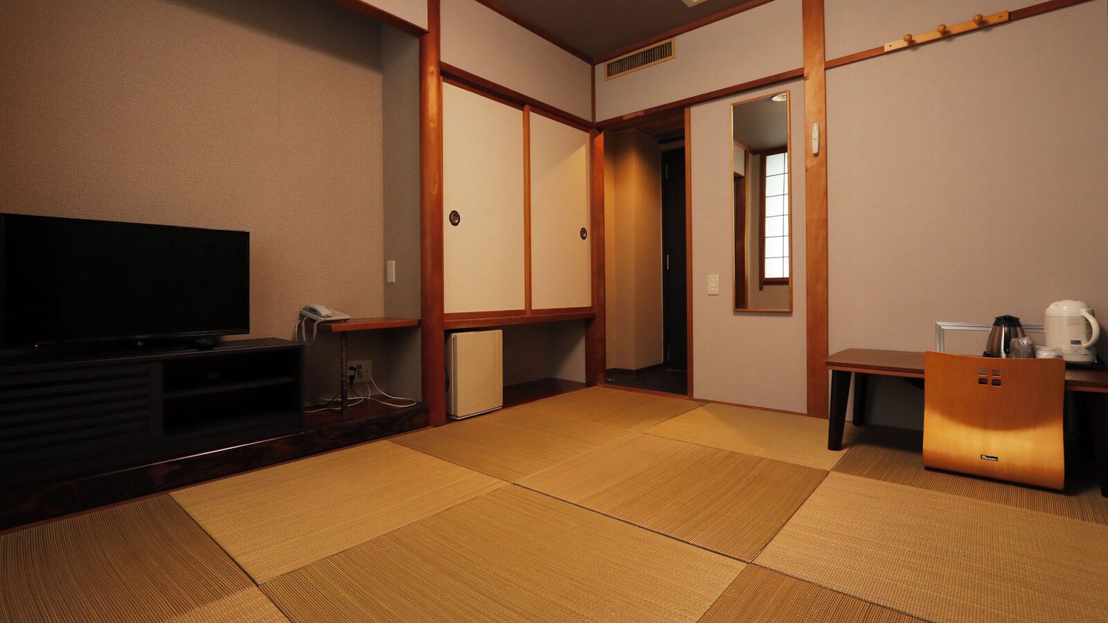 A traditional Japanese room with tatami flooring, a television, a small refrigerator, and a table with a kettle and cups.