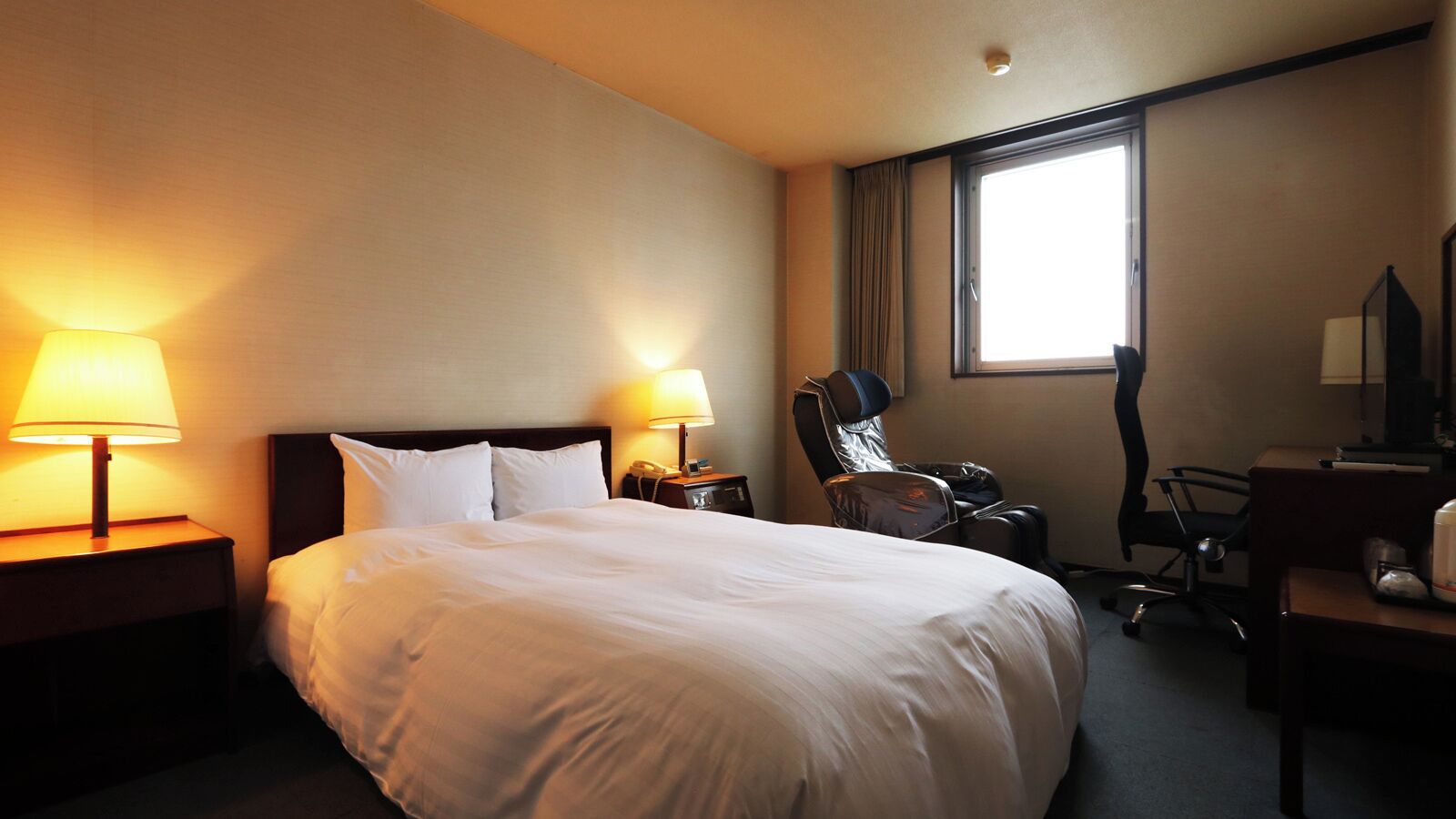 A hotel room with a large bed, two bedside lamps, a massage chair, a desk with a chair, and a window.