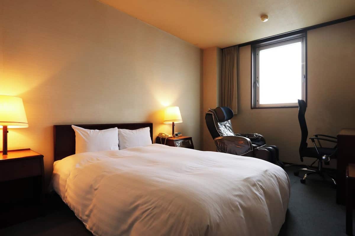 A hotel room with a large bed, two bedside lamps, a massage chair, a desk with a chair, and a window.