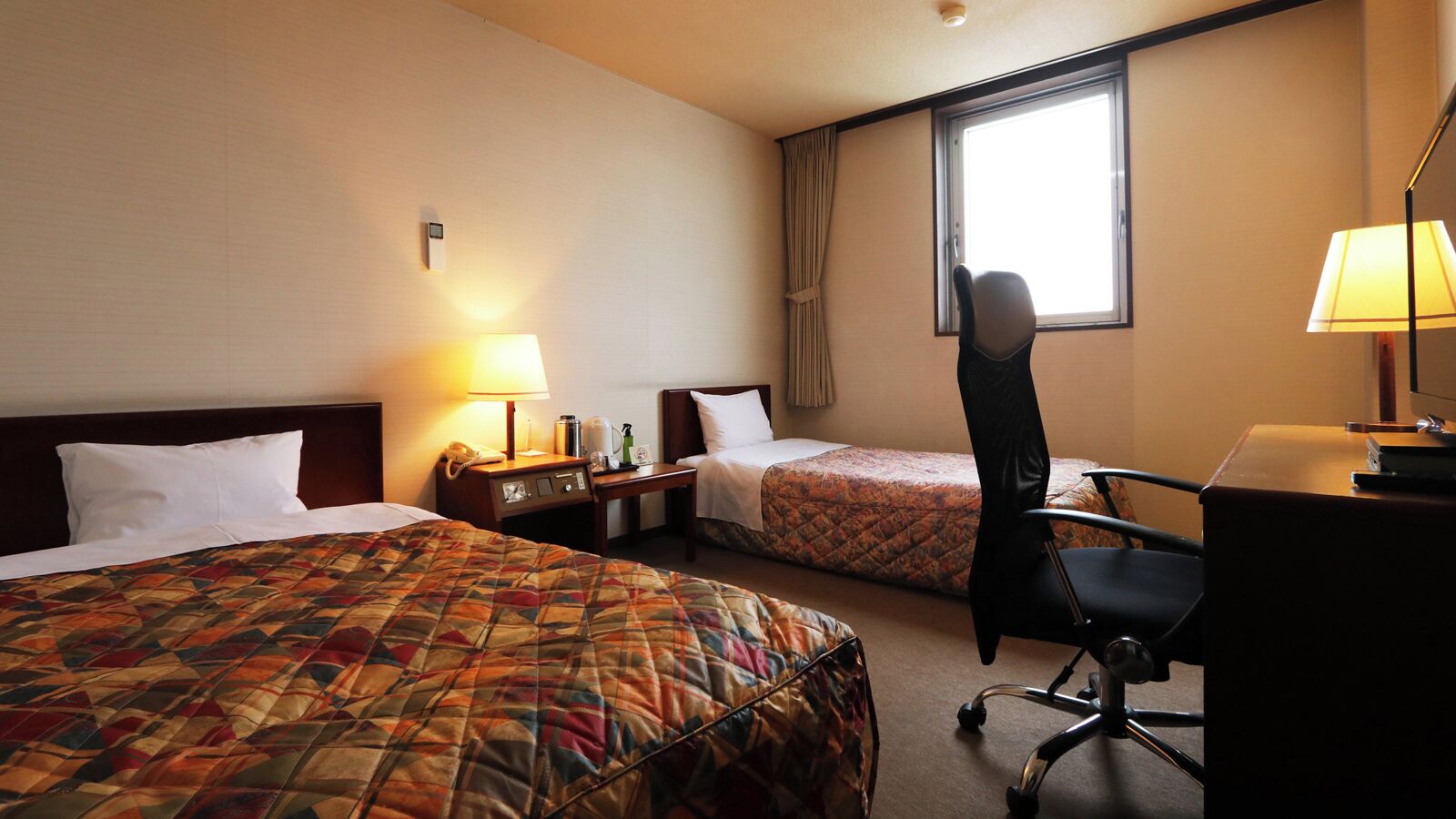 A hotel room with two beds, a desk, a chair, a window, and a lamp.