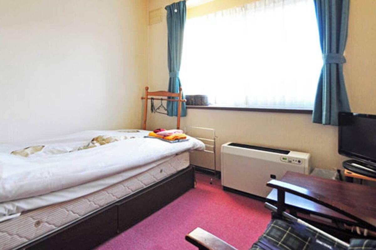 A single bed with a mattress, a bedside table, a heater, a TV, and a window with curtains.