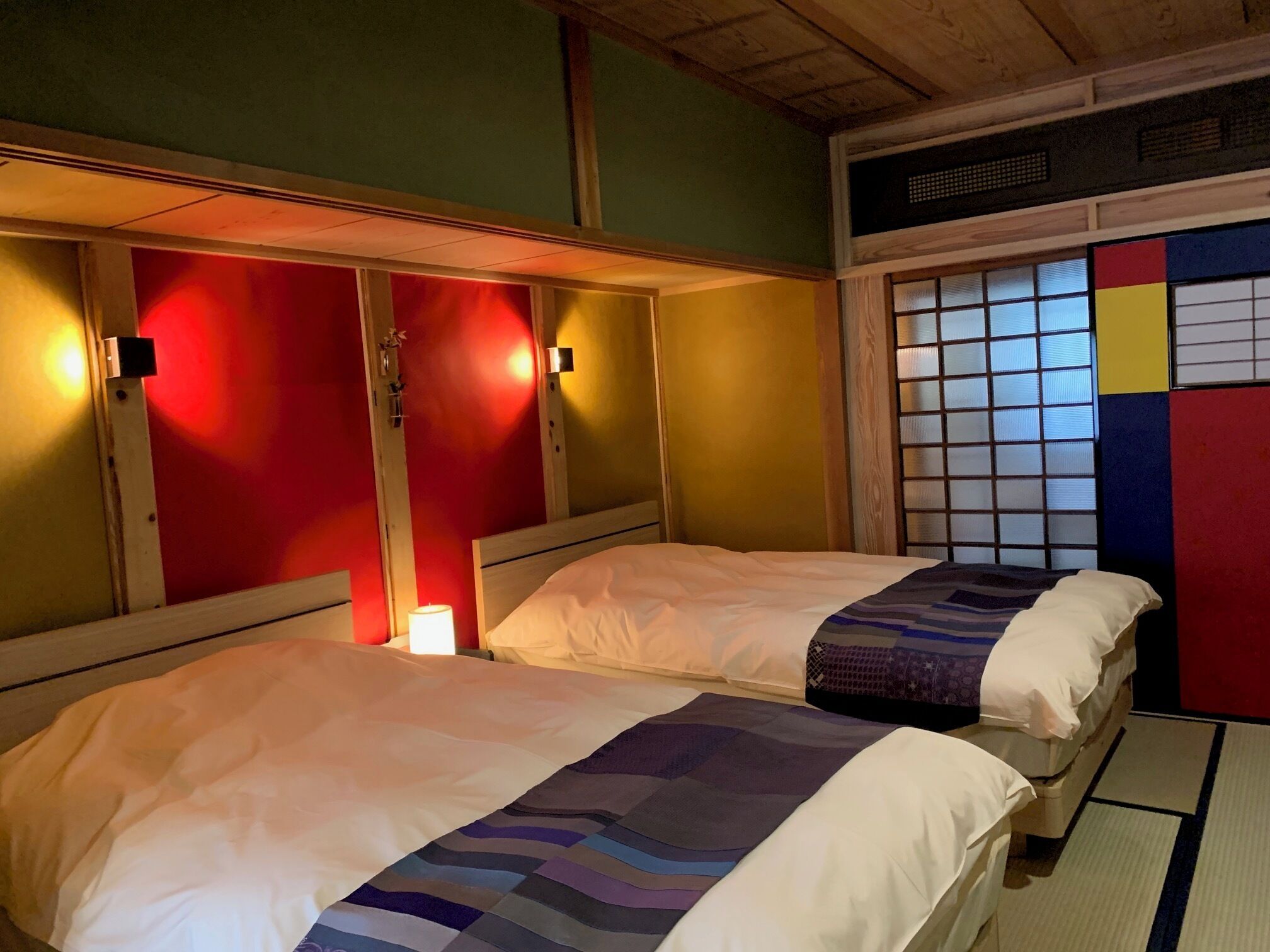 A room with two beds, a window with a grid pattern, and a wall with a colorful panel.