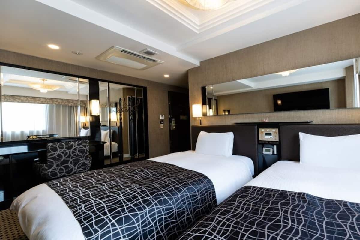 Ein Hotelzimmer mit zwei Betten, einem großen Spiegel, einem Schreibtisch mit Lampe und Blick auf die Hotelhalle.
