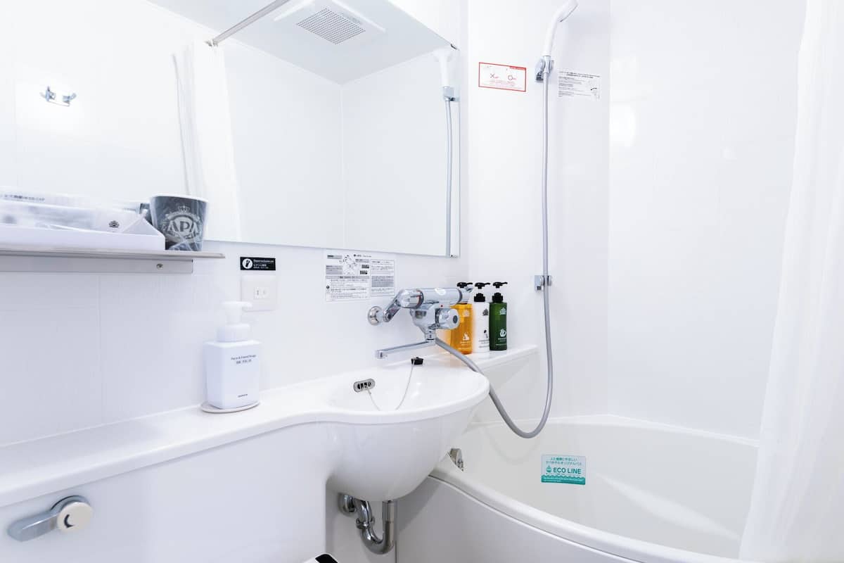 Ein modernes Badezimmer mit Waschbecken, Spiegel, Dusche und Toilette. Auf dem Waschbecken stehen Pflegeprodukte und ein Schild, das ein umweltfreundliches Wassersystem kennzeichnet.
