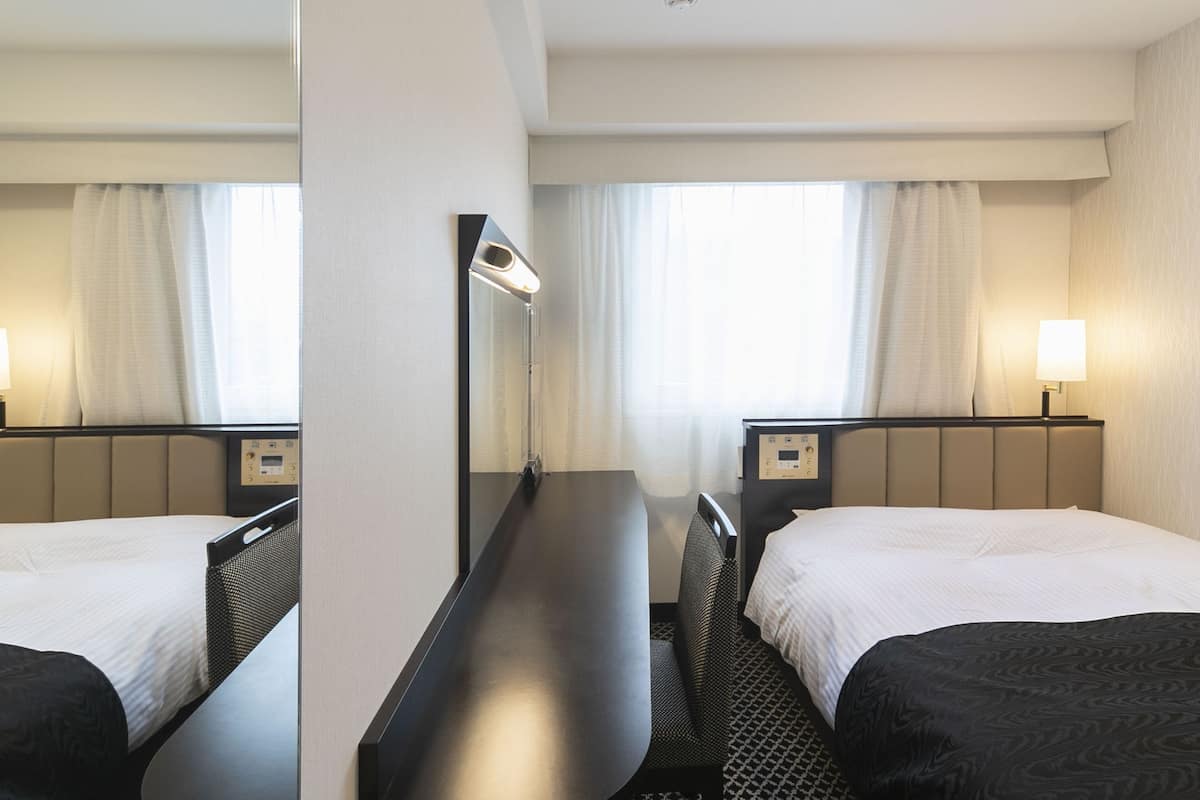 Ein Hotelzimmer mit zwei Betten, einem Schreibtisch, einem Stuhl, einer Lampe und einem fenster mit Vorhängen.