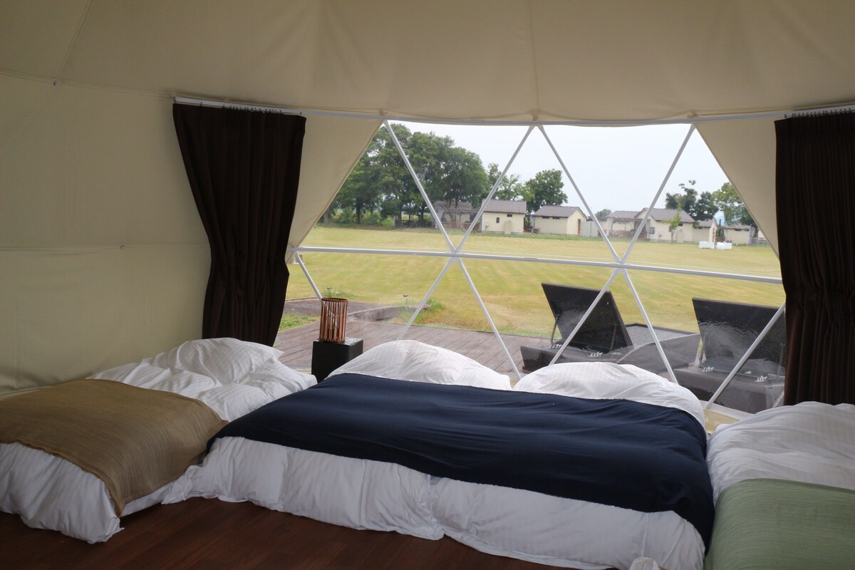 A tent with a bed, a chair, and a view of a grassy field with buildings in the distance.