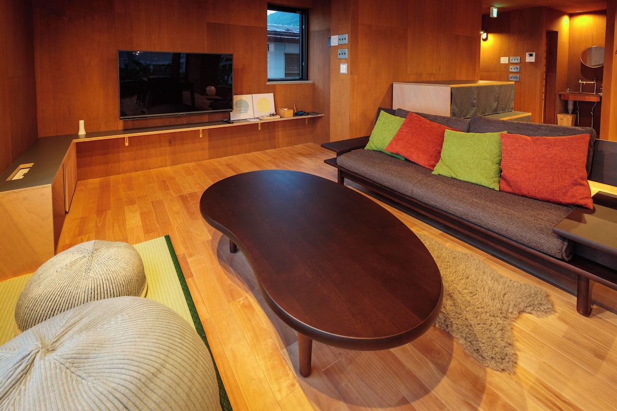 A modern living room with a wooden floor, a sofa with colorful cushions, a flat-screen TV, and a wooden coffee table.