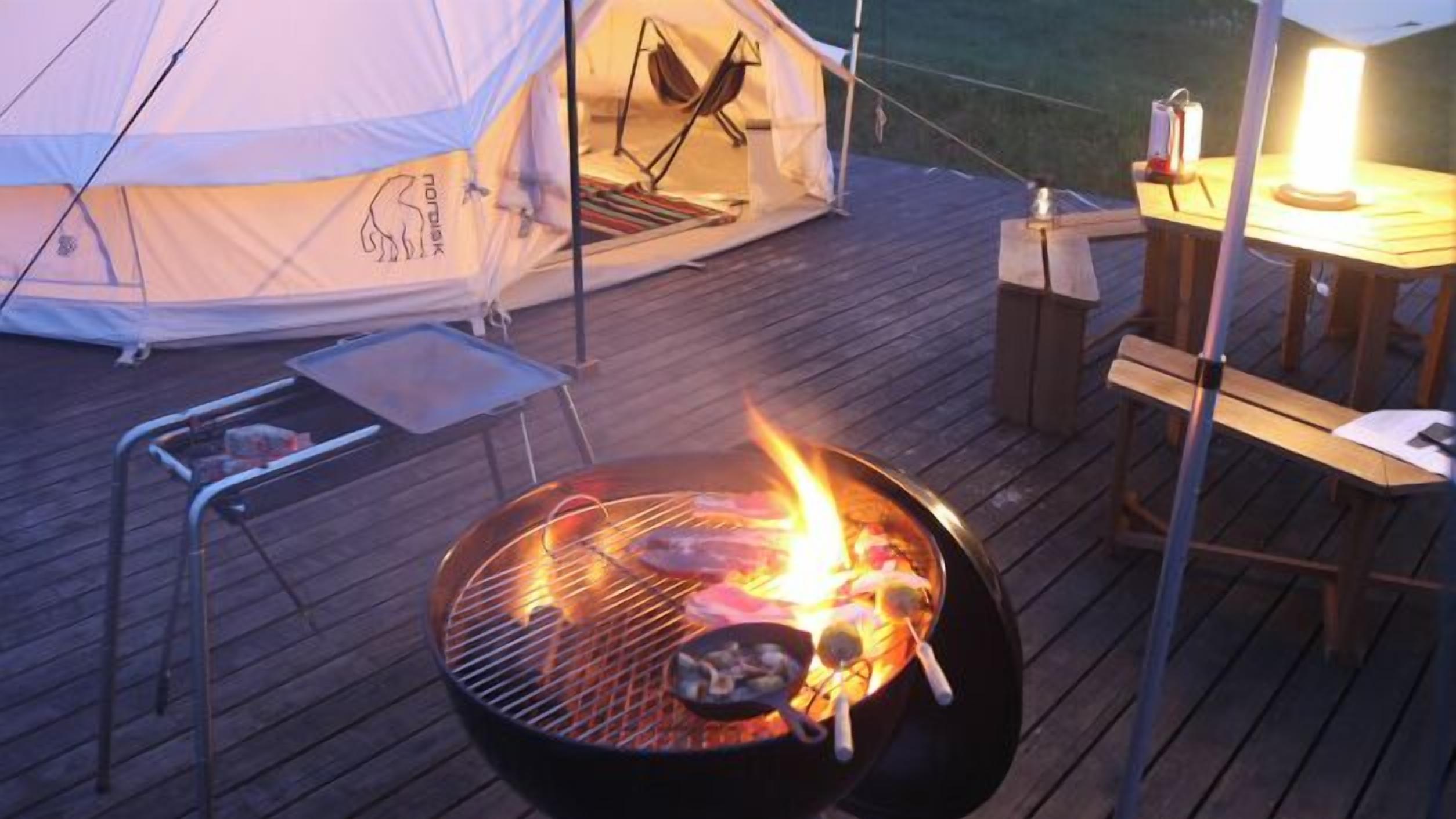 A tent with a sleeping area, a grill with food, and a table with a lamp.