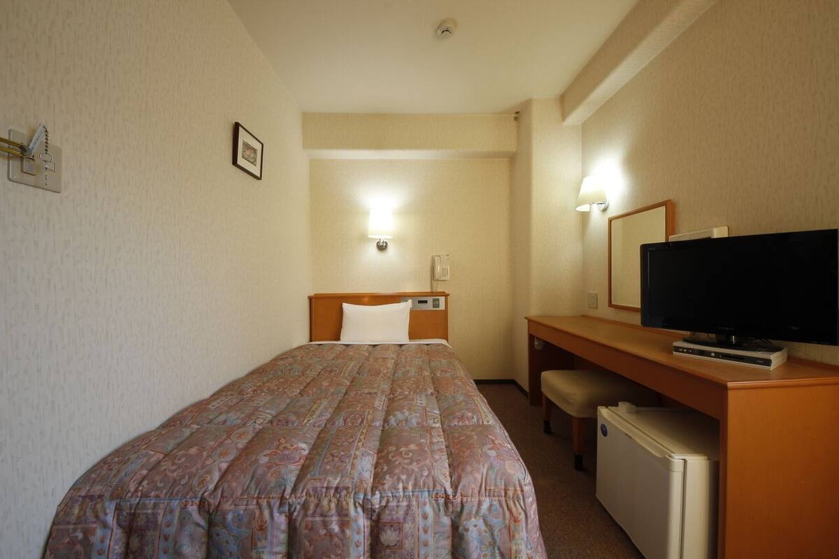 Habitación de hotel con una cama, un escritorio con televisor, un refrigerador pequeño y una lámpara fijada a la pared.