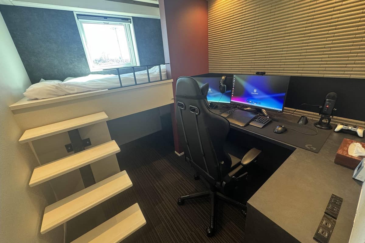 A compact hotel room with a bed, a desk with a computer, a chair, and a window with blinds.