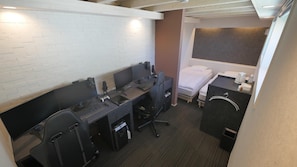 Ein kleines Hotelzimmer mit weißer Backsteinwand, einem Einzelbett, einem Schreibtisch mit zwei Computern und einem Nachttisch.