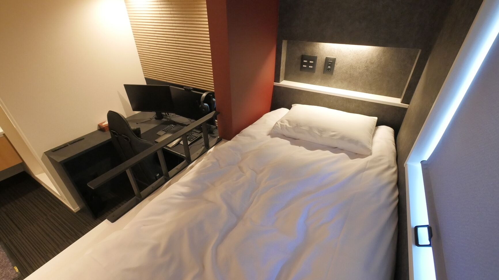 A hotel room with a bed, a desk with a computer, and a control panel on the wall.