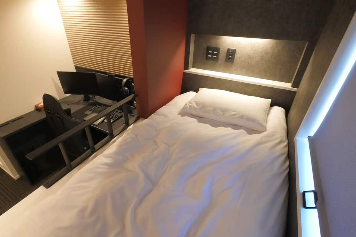 A hotel room with a bed, a desk with a computer, and a control panel on the wall.