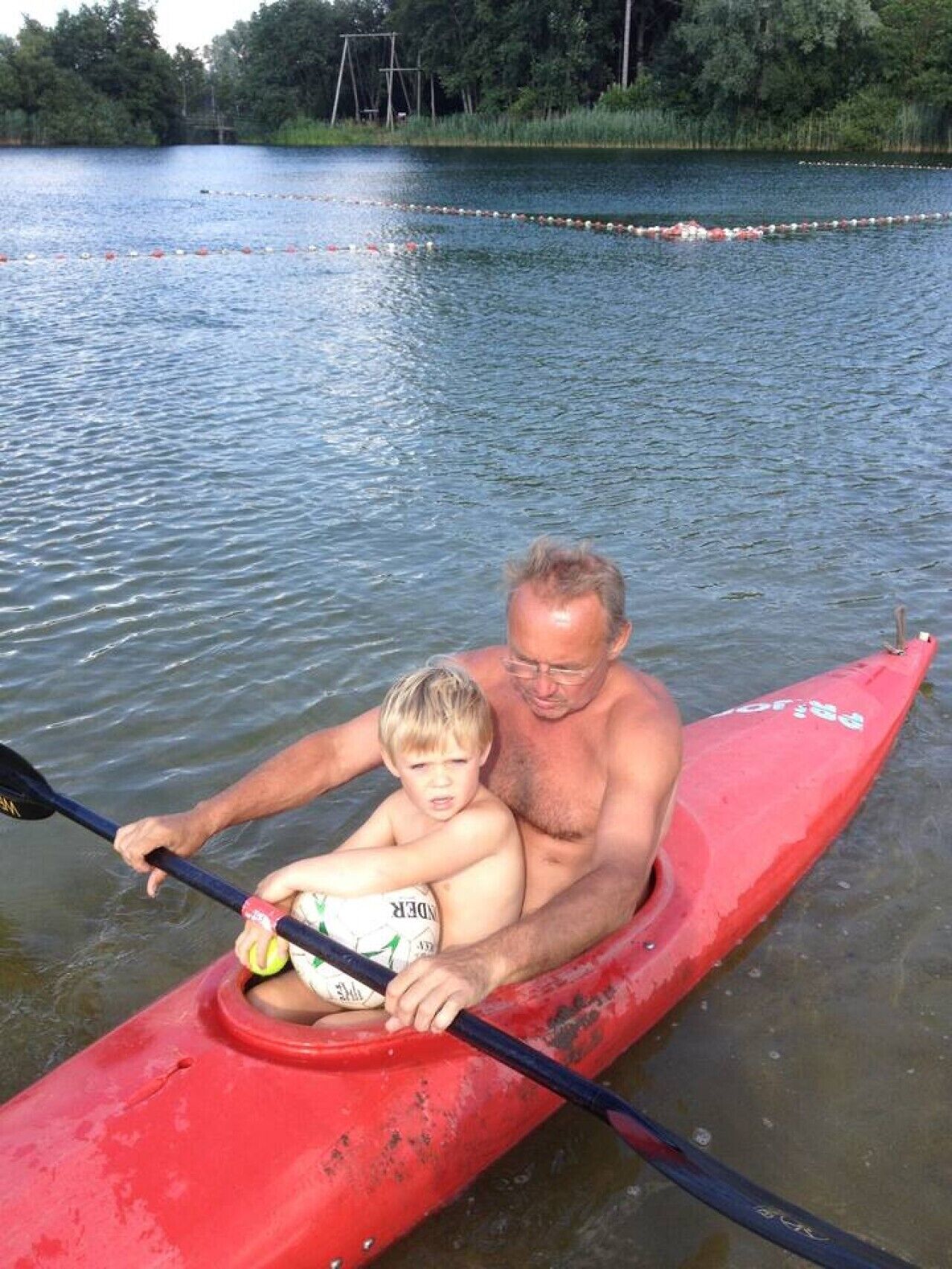 A man and a child in a red canoe on a calm lake with trees in the background.
