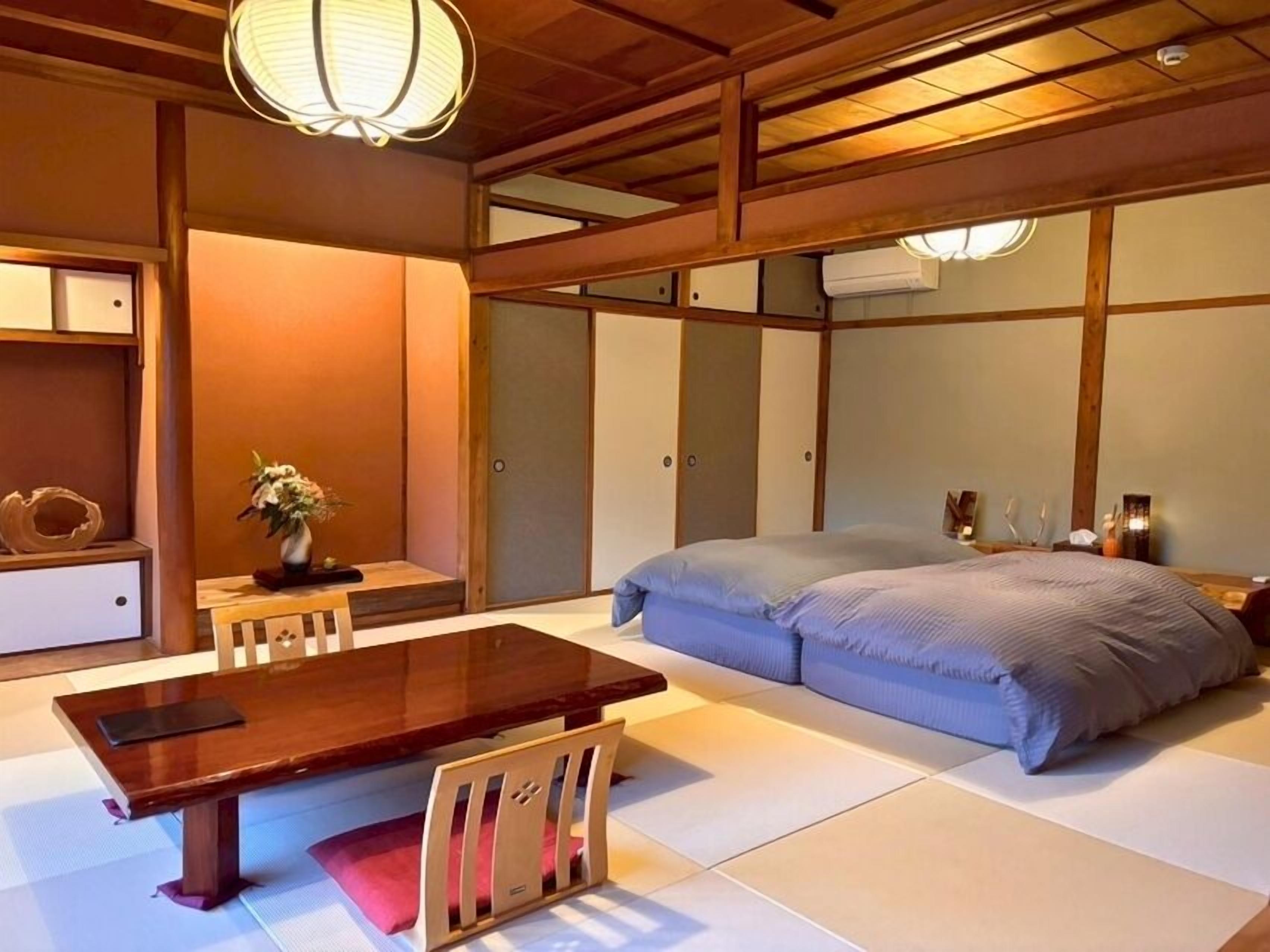 Um quarto tradicional em estilo japonês, com piso de tatame, uma mesa de madeira baixa, uma cama com roupa de cama azul e um lanterna de papel pendurada.
