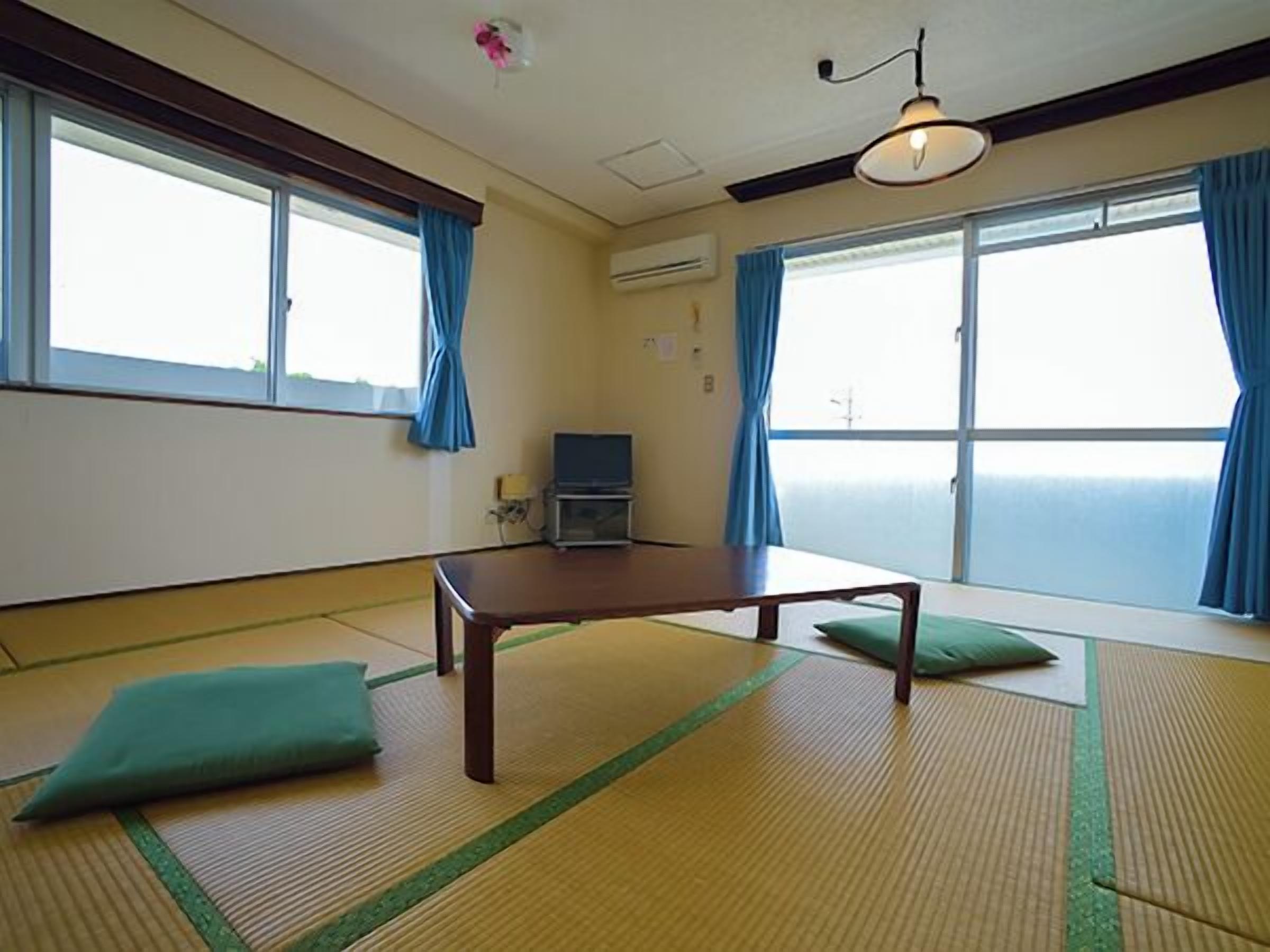 Ein Zimmer mit Tatami-Matten, einem niedrigen Tisch, einem Fernseher und großen Fenstern mit blauen Vorhängen.