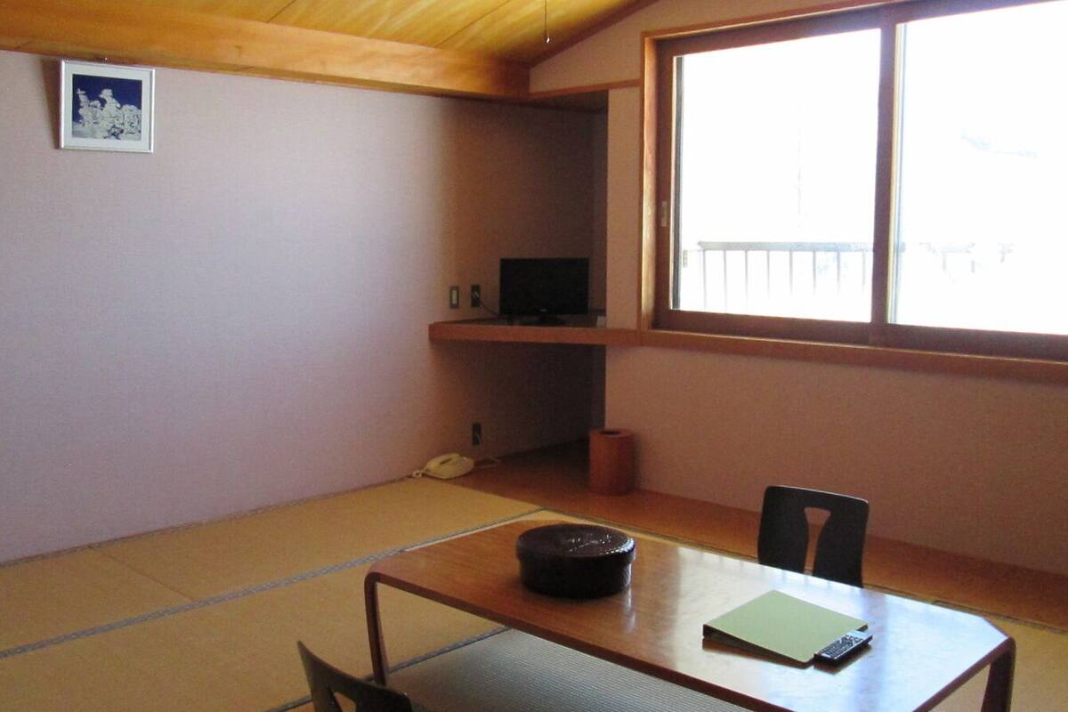 A room with tatami flooring, a low table with a cushion, a chair, a TV on a shelf, and a window with a view.