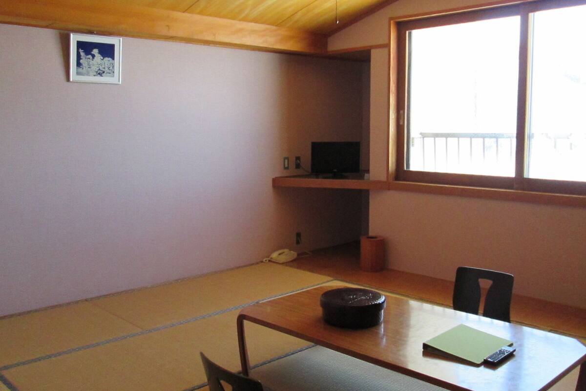 A room with tatami flooring, a low table with a chair, a television on a shelf, and a window with a view.