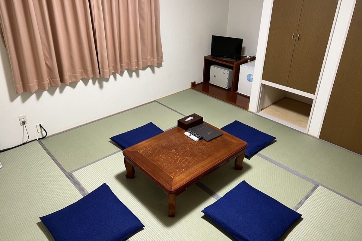 A tatami mat room with a wooden table, blue cushions, a television, and a small cabinet.