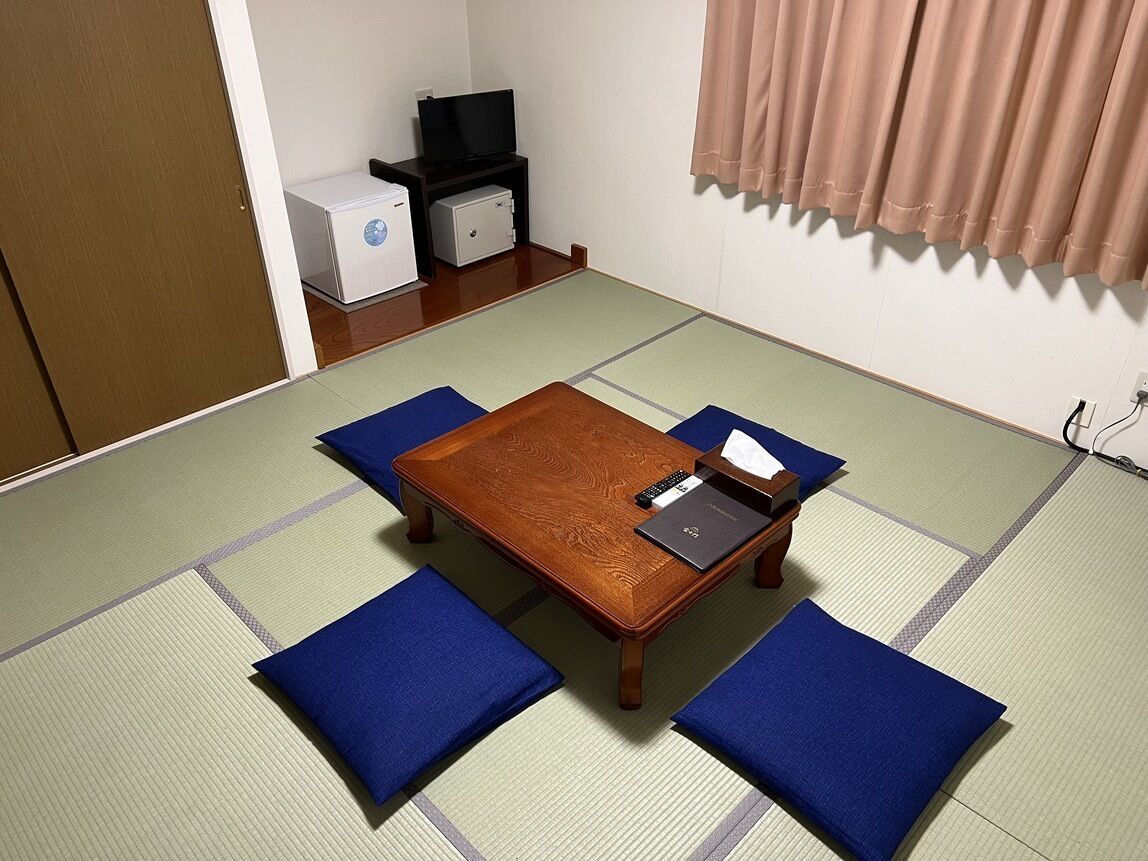 Um quarto com esteiras de tatame, uma mesa de madeira, almofadas azuis, uma televisão e um pequeno frigorífico.