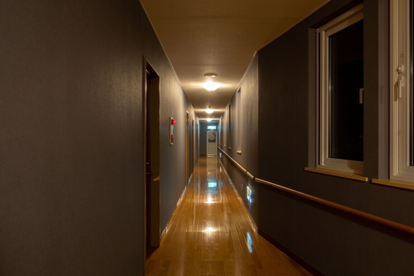 A long, narrow hallway with wooden flooring, doors on both sides, and a window on the right.