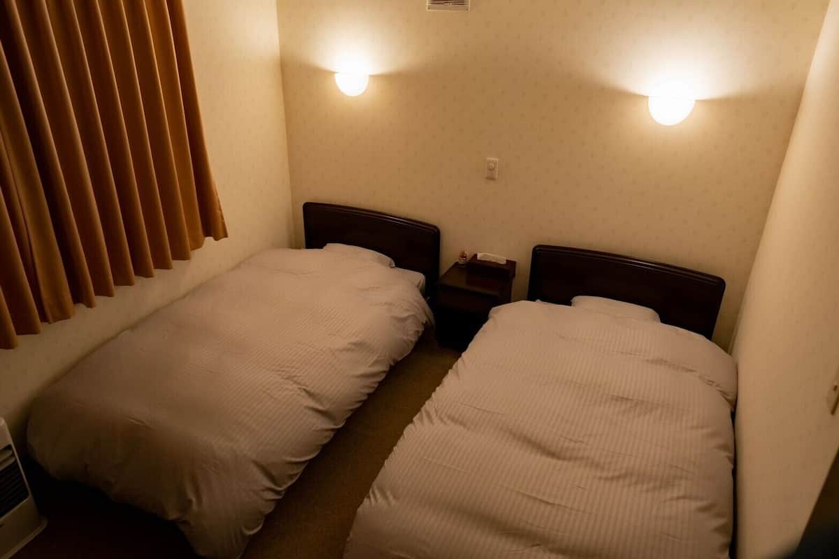 A small, dimly lit room with two single beds, a nightstand, and a wall-mounted light fixture.