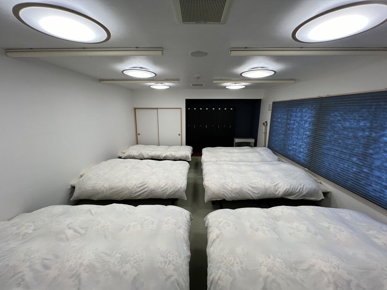 Quarto com quatro camas dispostas em linha, uma porta e uma janela com persianas.