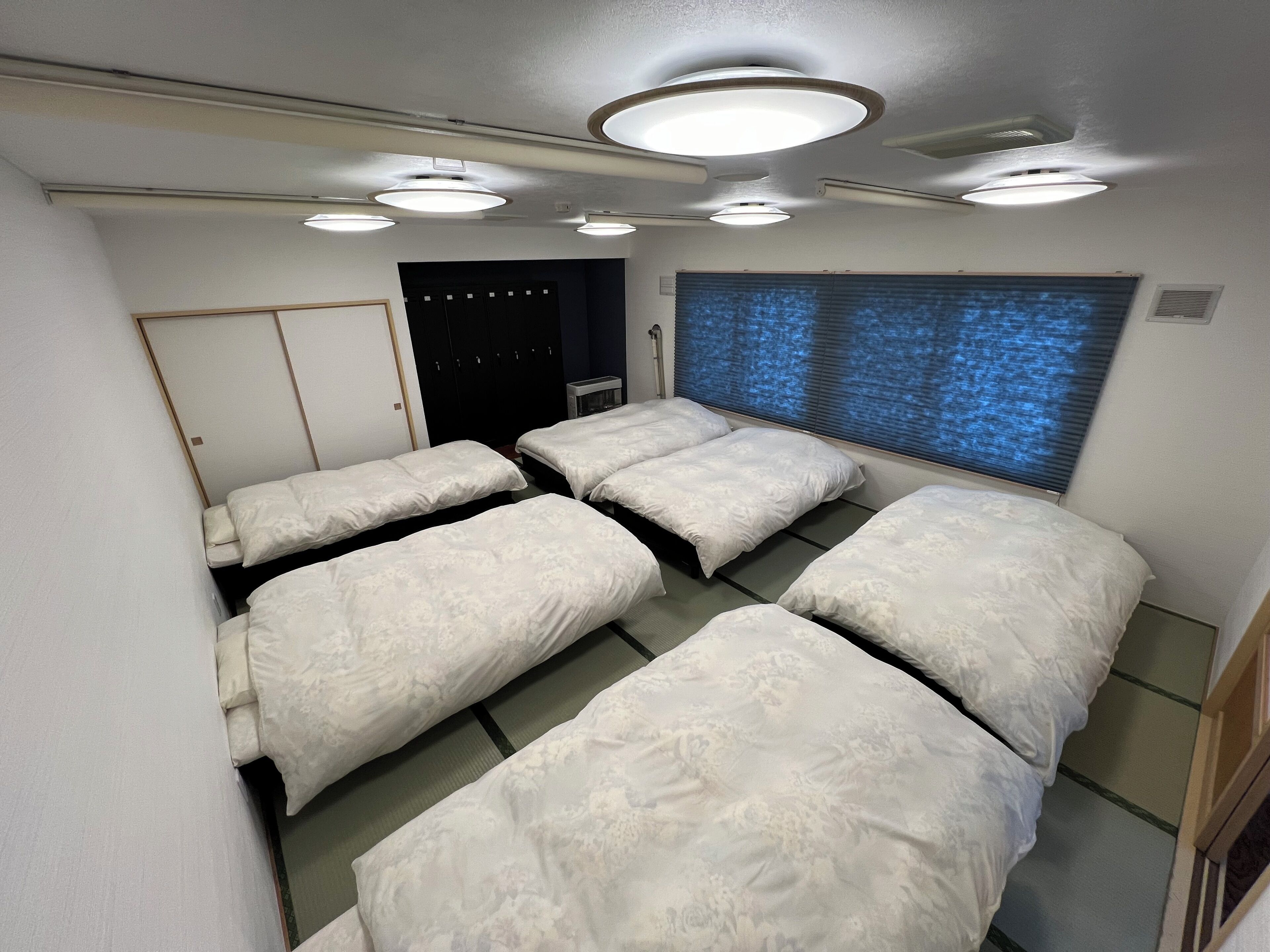 Quarto de hotel com quatro camas, um guarda-roupa, janela com persianas e iluminação no teto.