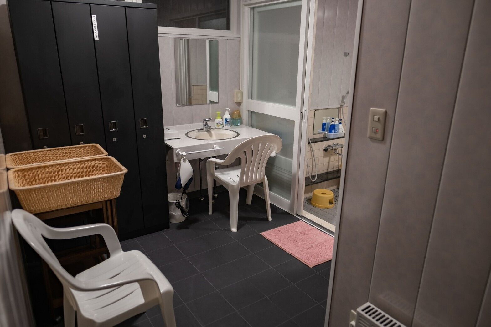 Een badkamer met een wastafel, spiegel, witte stoelen, rieten manden en een douchegedeelte.