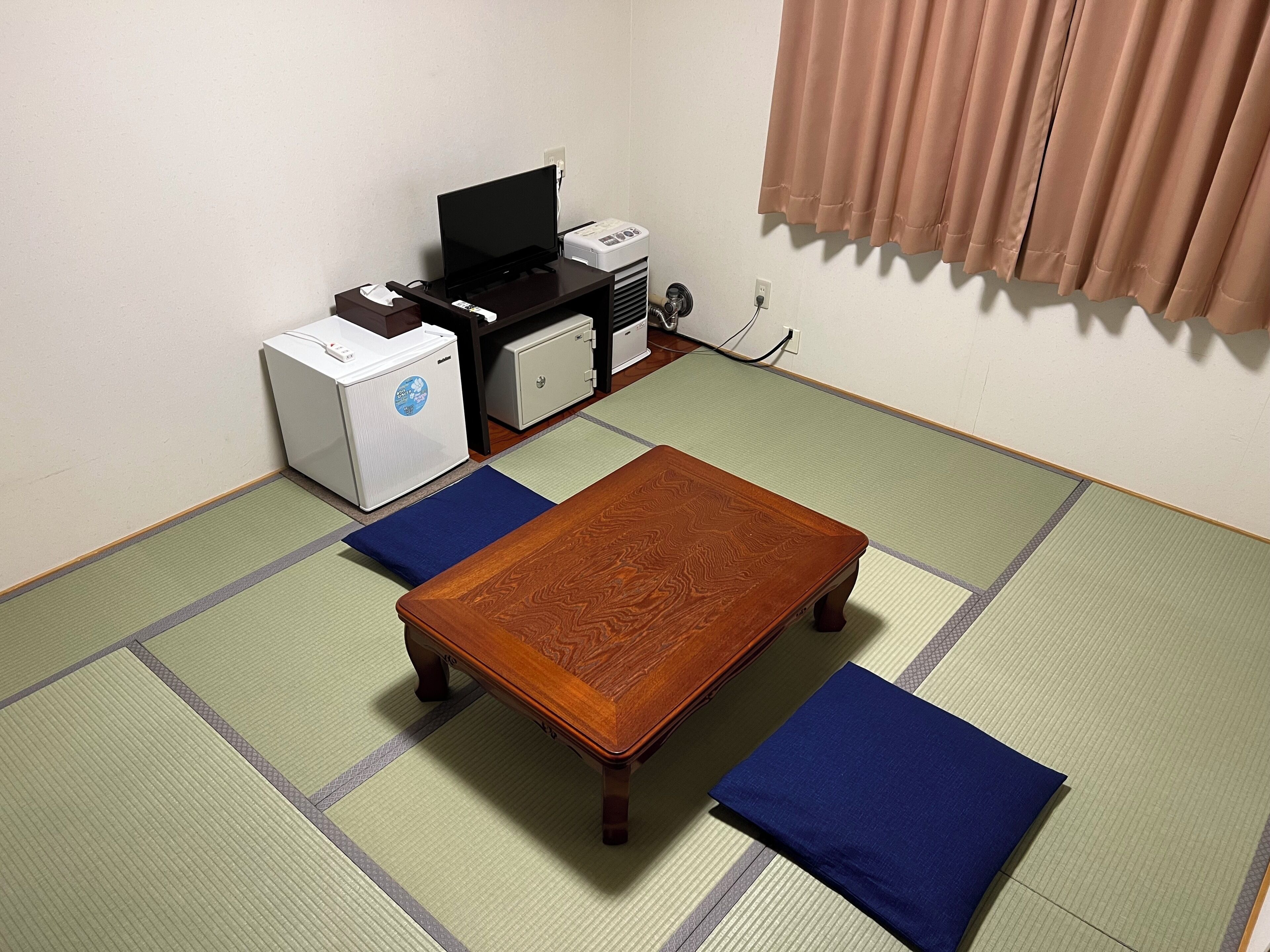 Ein Zimmer mit Tatami-Böden, einem Holztisch mit Kissen, einem Schreibtisch mit Computer und einem kleinen Kühlschrank.