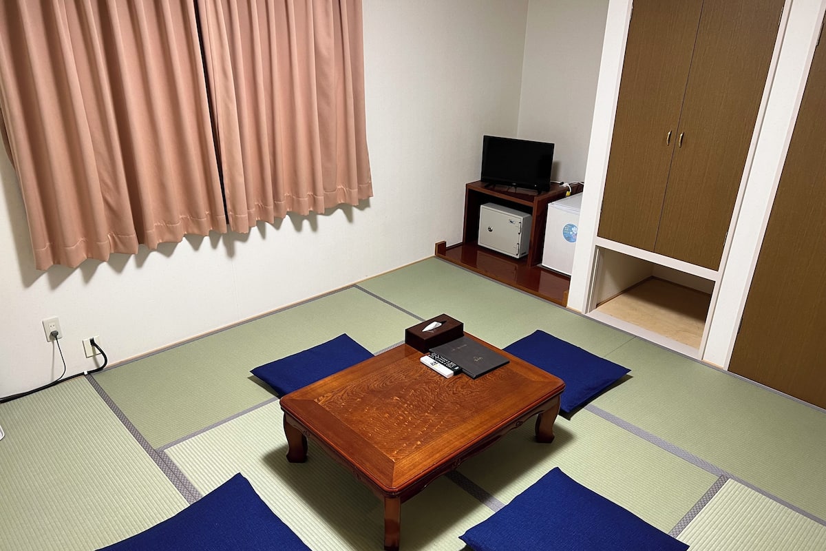 A traditional tatami mat room with a wooden table, blue cushions, a television on a stand, and a wardrobe.