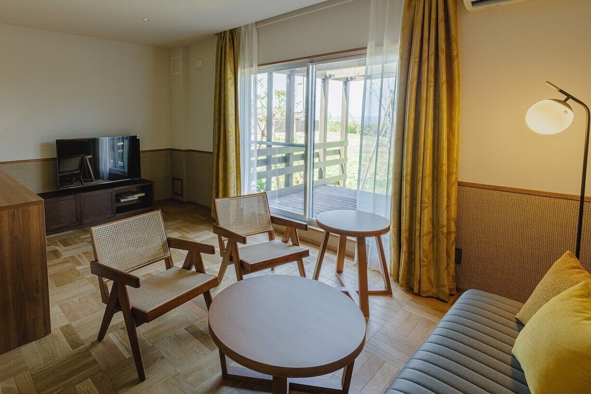 Quarto de hotel com sofá, mesa redonda, duas cadeiras, televisão e varanda com cortinas.