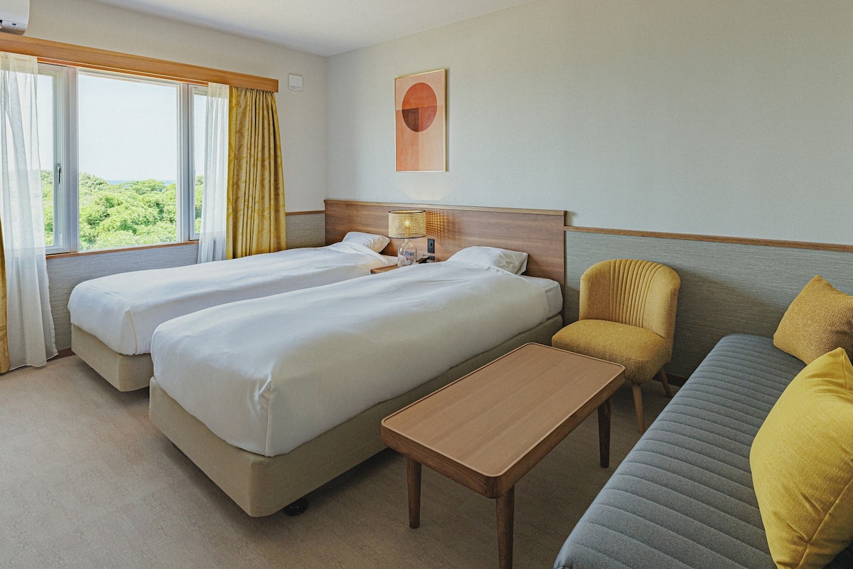 Quarto de hotel com duas camas, um sofá, uma mesa pequena e uma janela com cortinas.