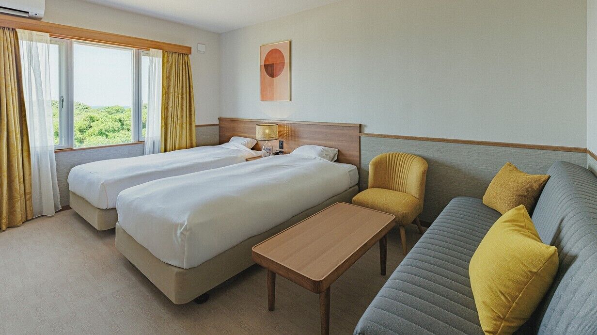 Ein Hotelzimmer mit zwei Betten, einem Schlafsofa, einem kleinen Tisch und einem Fenster mit Vorhängen.