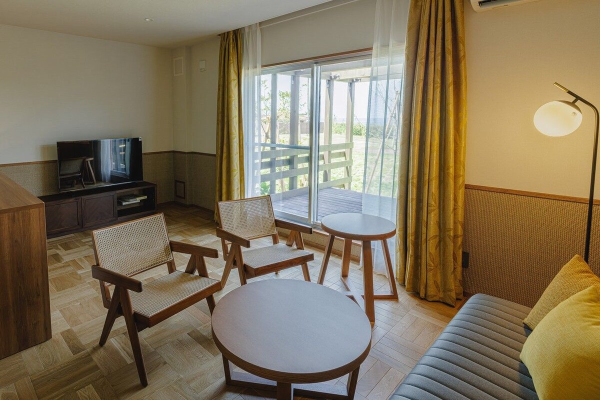 Quarto de hotel com sofá, mesa redonda, duas cadeiras, televisão e varanda com cortinas.