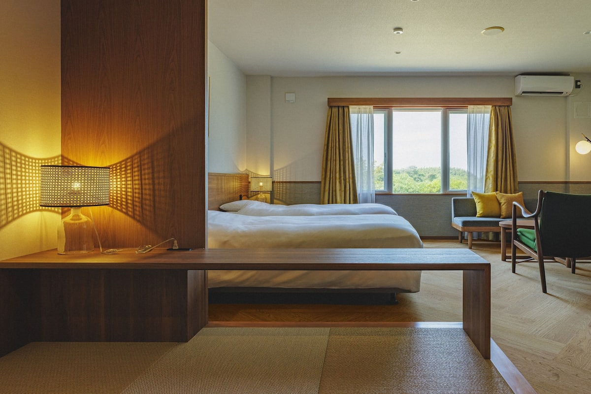 Quarto de hotel com cama, escrivaninha, cadeira, televisão e janela com cortinas.