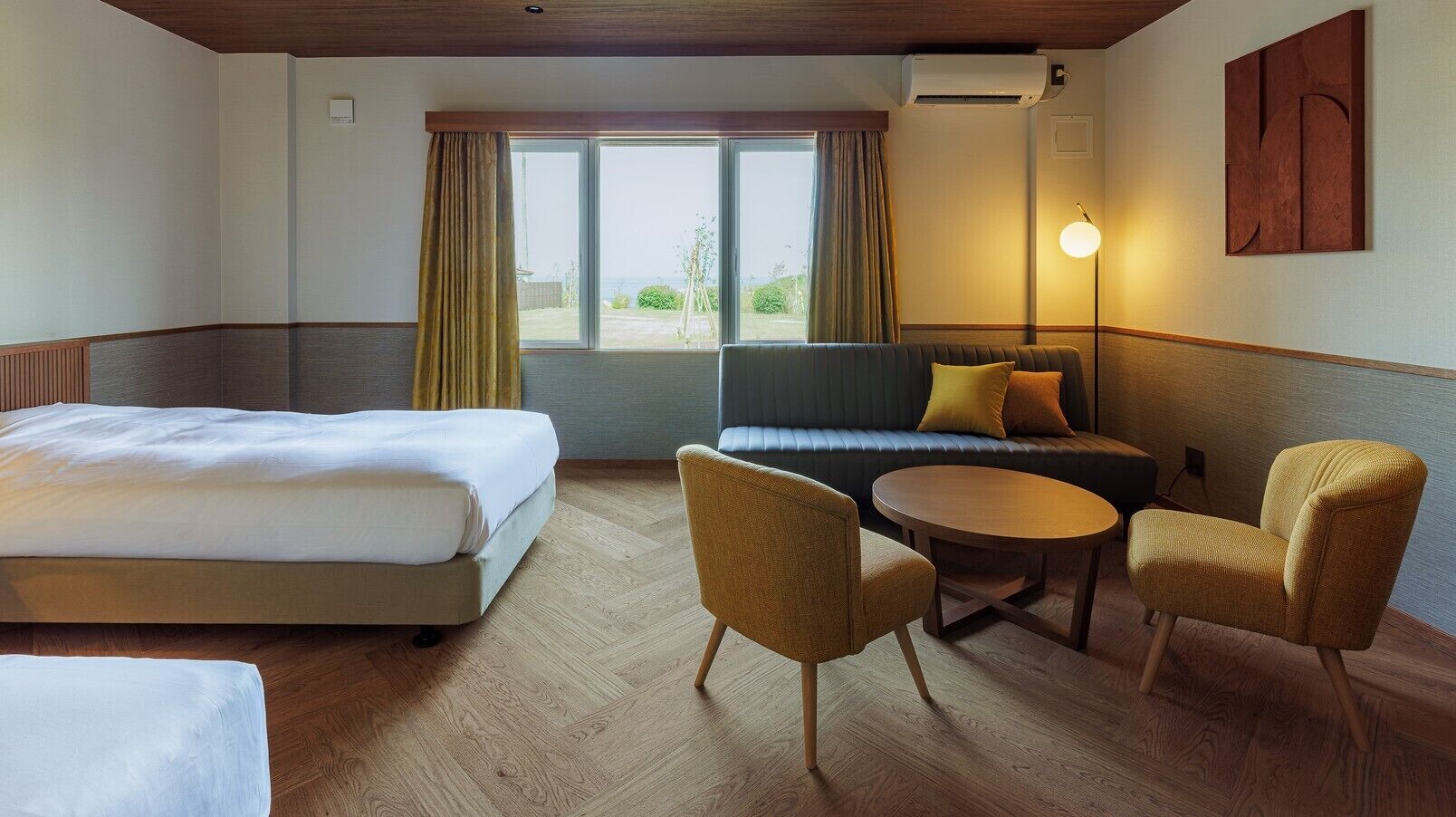Ein Hotelzimmer mit einem Bett, einem Sofa, zwei Sesseln, einem kleinen Tisch und Blick durch ein großes Fenster.