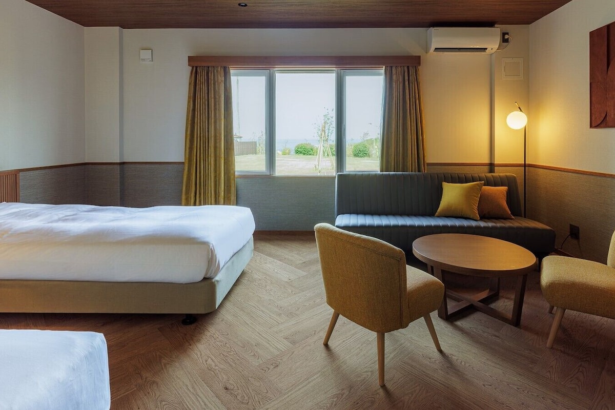 Quarto de hotel com uma cama, um sofá, duas poltronas, uma mesa pequena e vista pela grande janela.